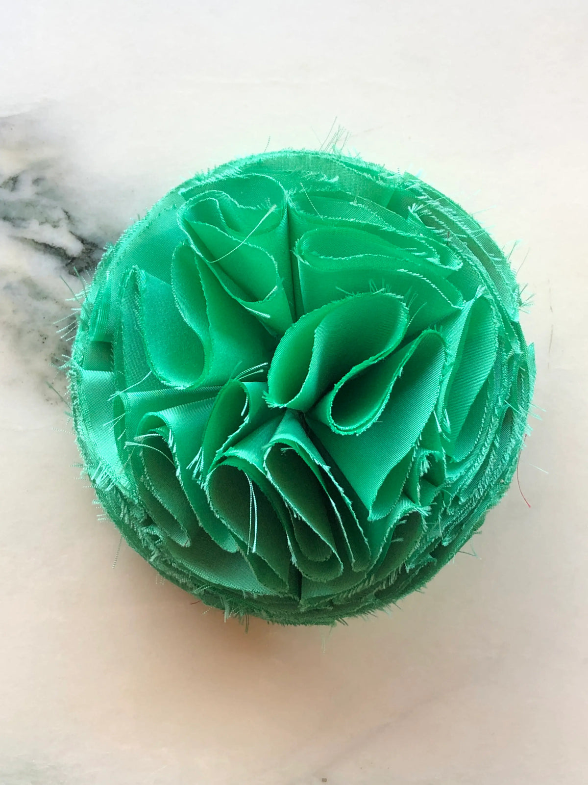 A Broochella Men's Green Pom Pon small fabric flower brooch with layered raw-edge petals, photographed on a white marble background with soft grey veining.