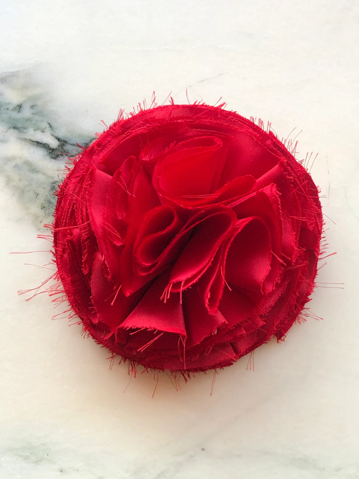 A Broochella Men's Red Pom Pon small fabric flower brooch with layered raw-edge petals, photographed on a white marble background with soft grey veining.
