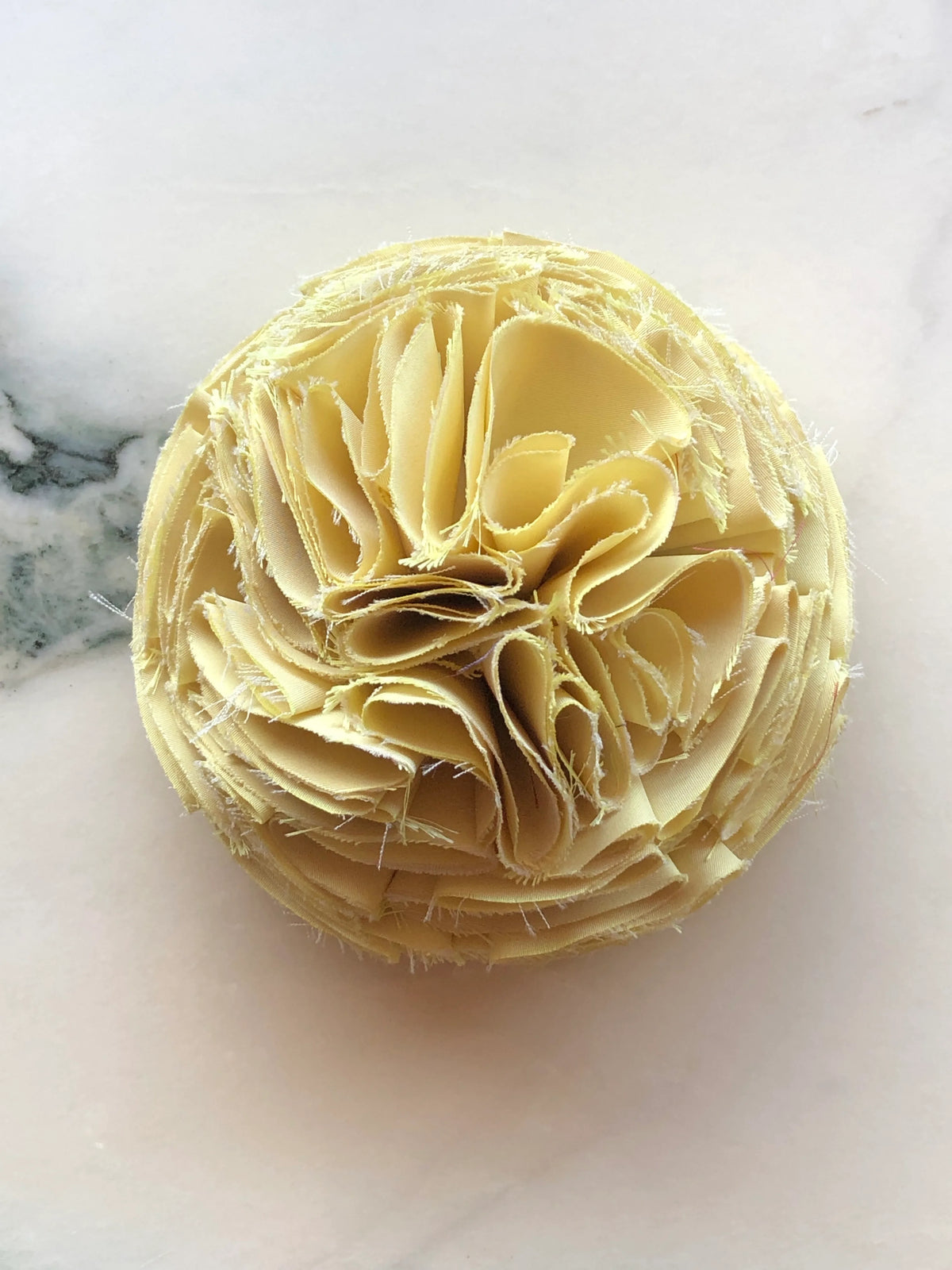 Men's Broochella Yellow Pom Pon small fabric flower brooch with layered raw-edge petals, photographed on a white marble background with soft grey veining.